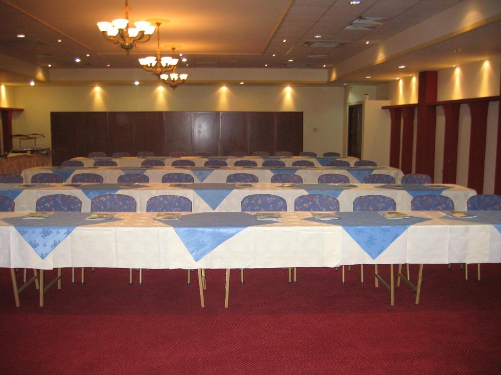 Konferenču zāle ar rindu galdiem un krēsliem, dekorēta ar zilu un baltu galda veļu. Telpa ir labi apgaismota un gatava sanāksmēm vai pasākumiem.