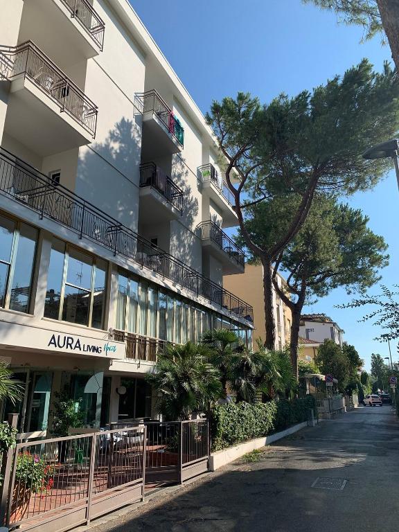 Mūsdienīga ēka ar balkoniem un plašu logu, kurā redzama zaļa ainava, palmām un augstiem kokiem. Gaismas pilna iela ar zilu debesu fonu.