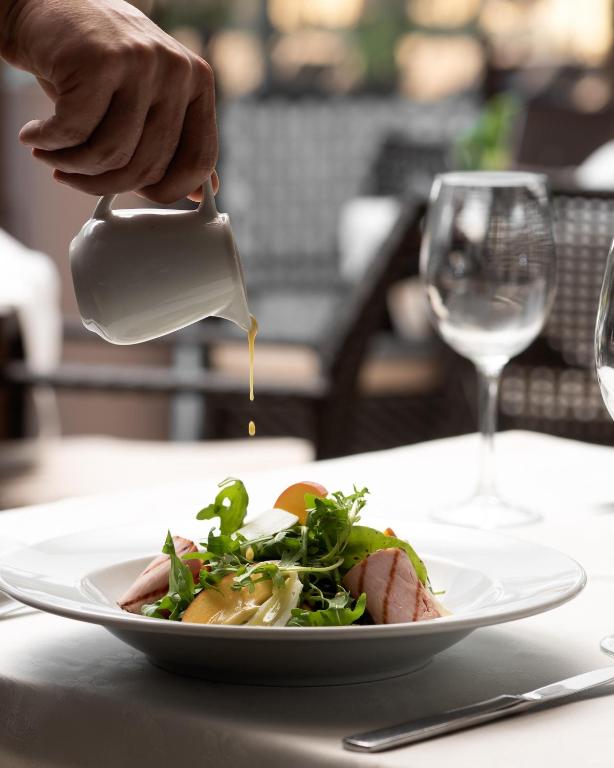 Svaigas salāti tiek apēsti ar mērci, kas tiek uzlieta no baltas krāsas mērces trauka. Fons ir elegants restorāns ar stikla glāzēm un mīkstām mēbelēm.