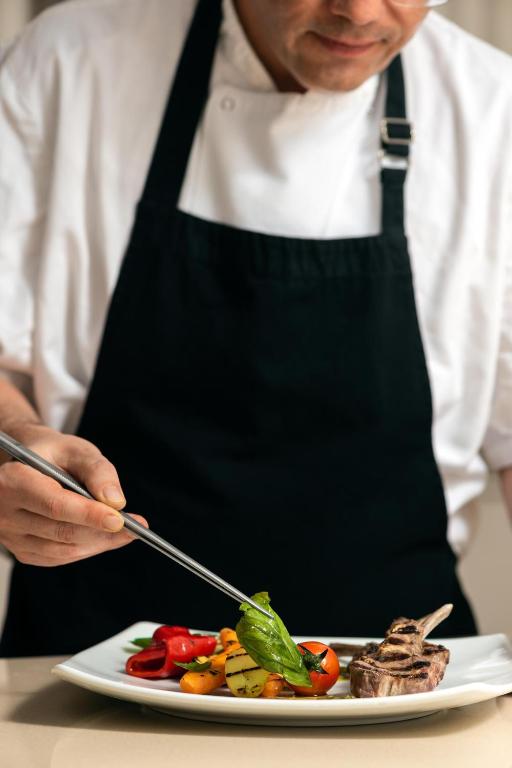 Šefpavārs ar šķērsgriezuma rīkiem dekorē šķīvi ar grilētiem dārzeņiem un gaļas ēdienu. Šis attēls izceļ kulinārijas mākslu un uzmanību detaļām.