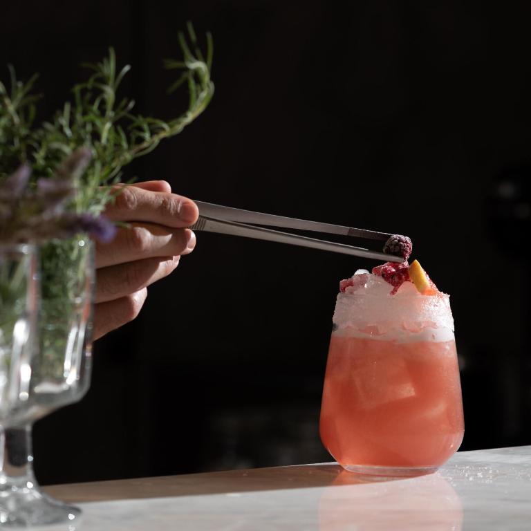 Kokteilis ar ledus un augļiem, ko pasniedz ar pinceti. Fons ir tumšs, akcentējot dzēriena krāsas un tekstūru.