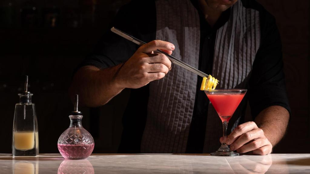 Barmenis pievieno ananāsus pie kokteiļa, izmantojot ēdiena kociņu, kamēr uz galda ir dažādas pudeles ar dzērieniem.