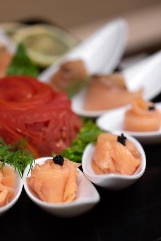 Svaigi pasniegts kūpināts lasis uz baltām karotēm, dekorēts ar melnajām ikriem un zaļumiem. Fonā redzami dažādi zivju ēdieni un garnējums.