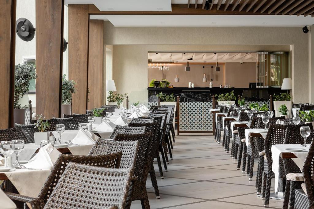 Mūsdienīgs restorāns ar elegantiem galdautiem un vītņotiem krēsliem, kas izveidots gaišā un komfortablā vidē. Interjerā redzami augi un moderns bārs.