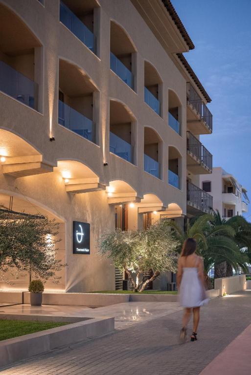 Elegants viesnīcas eksterjers ar mūsdienīgu dizainu un apgaismojumu, kurā sieviete staigā gar gājēju celiņu. Viesnīca ir ieskauta zaļumiem un palmām, radot relaksējošu atmosfēru.