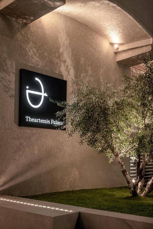 Mūsdienīgs viesnīcas "Theartemis Palace" logo, kas izceļas uz vienkāršas sienas fona, apstādīts ar zaļu olīvkoku.