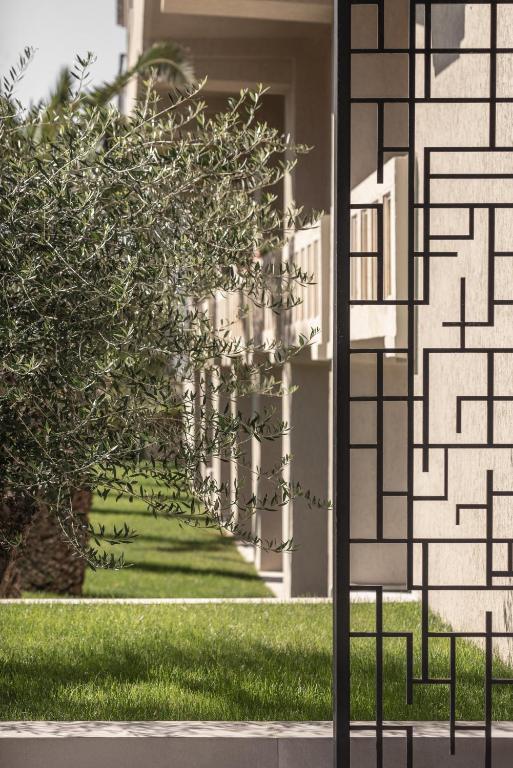 Dizains ar mūsdienīgu metāla žogu, kas atdala zaļo zālienu un olīvu koku no modernās arhitektūras fona.