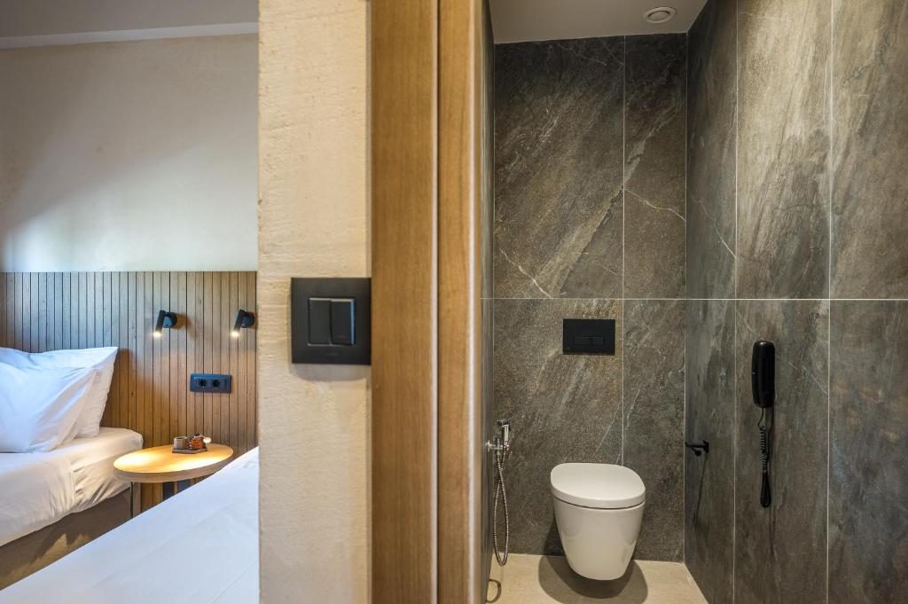 Moderns viesnīcas numurs ar gultu un vannas istabu, kas ir atdalīta ar sienu. Gultā ir balti spilveni, un blakus atrodas mazs galdiņš. Vannas istaba ir apdarināta ar akmens flīzēm un aprīkota ar mūsdienīgu tualeti un telefonu.