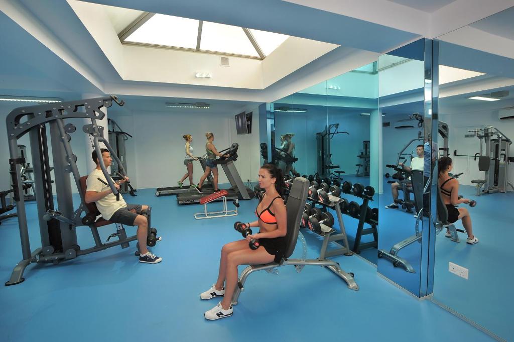 Mūsdienīga sporta zāle ar dažādiem trenažieriem un svaru komplektiem, kur divi cilvēki veic vingrinājumus. Telpa ir gaiša un plaša ar spoguļiem un logu, kas nodrošina dabisko apgaismojumu.
