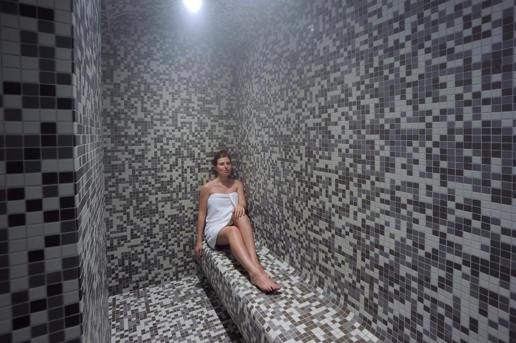 Sieviete baltā dvielī relaksējas modernā mozaīkas stila tvaika pirtī ar pelēkiem toņiem. Šī attēla atmosfēra atspoguļo mierīgu un komfortablu spa pieredzi.