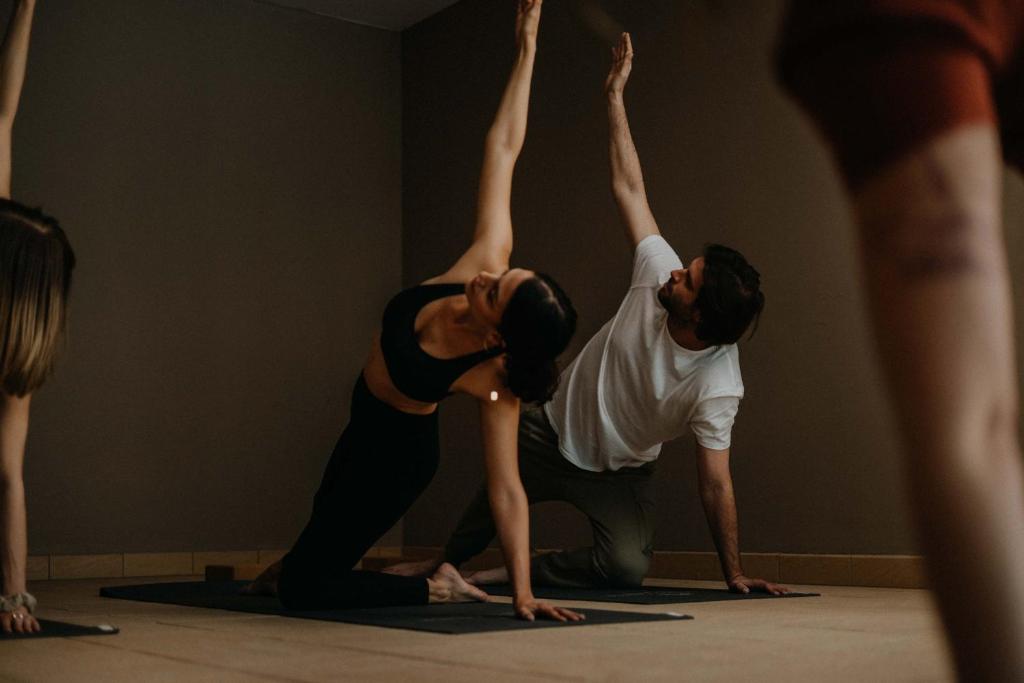 Joga nodarbība ar diviem cilvēkiem, kas veic pozas uz paklājiem. Telpa ir mierīga un apgaismota ar neitrālām krāsām.