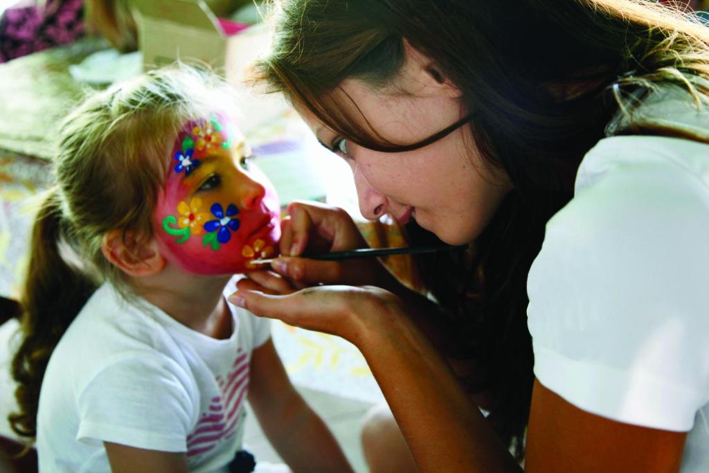 Bērns ar krāsotu seju saņem sejas krāsošanu no pieaugušā. Attēls atspoguļo jautru un radošu brīdi, kad tiek izveidotas krāsainas ziedu un citu rakstu detaļas.