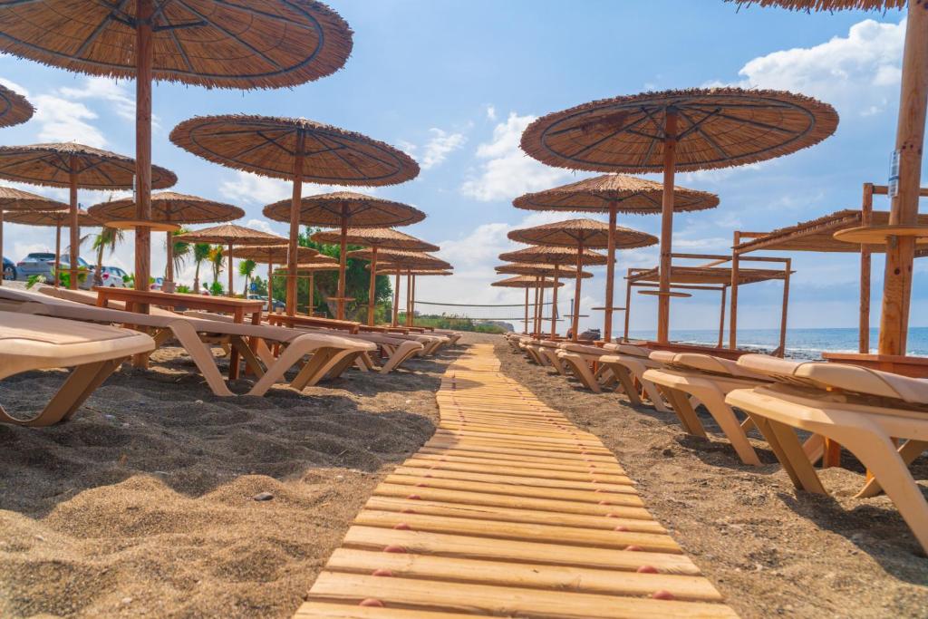 Pludmales ainava ar koka sauļošanās krēsliem un salmu jumtiem, kas izkārtoti pa smilšainu ceļu, kas ved uz jūru. Skats uz saulainu debesi un mierīgu jūras viļņu plūdumu.