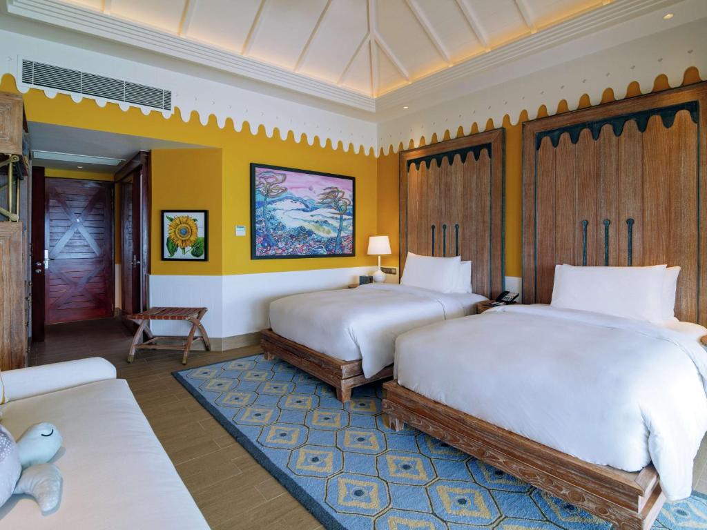 Mūsdienīga viesnīcas numura interjers ar divām gultām, gaišiem toņiem un krāsainu mākslas darbu uz sienas. Telpā ir ērta atpūtas zona un siltas krāsas akcenti.