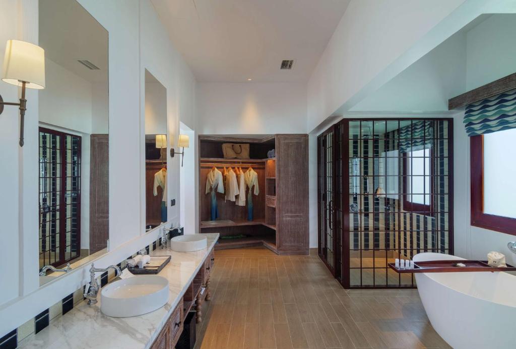 Mūsdienīgs vannas istabas interjers ar divām izlietnēm, lielu vannu un drēbju skapi, kas atrodas pie griestiem. Labi apgaismota telpa ar spoguļiem un elegantu dizainu.