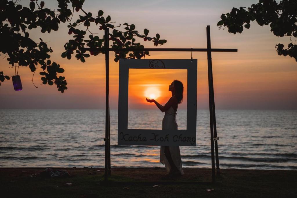 Silhouette of a woman holding the sun at sunset, framed by a decorative photo frame on the beach. The vibrant colors of the sky reflect a serene tropical atmosphere.