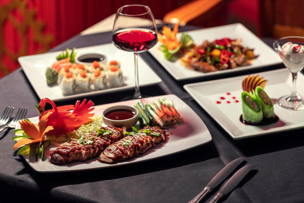 Elegants restorāna ēdienu komplekts ar dažādiem traukiem, tostarp grilētu gaļu, suši un desertiem, pasniegts ar glāzi sarkanvīna.