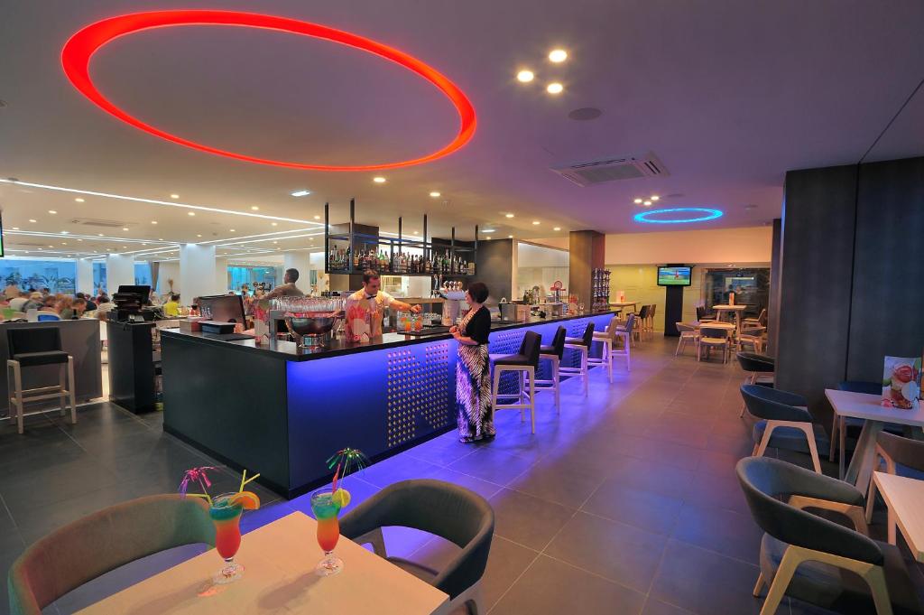 Moderns bārs ar krāsainu apgaismojumu un dzērieniem uz galda, kas radījis patīkamu atmosfēru. Klienti izbauda savus dzērienus un sarunas mērenā, stilīgā vidē.