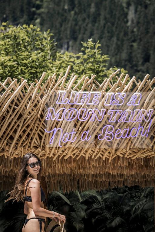 Dzīve ir kalns, ne pludmale! Sieviete ar brīvdienu apģērbu stāv zem bambusa nojumes, kas rotāta ar neona uzrakstu.
