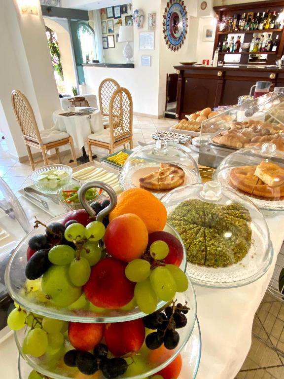 Bagātīgs brokastu galds ar svaigiem augļiem un gardām uzkodām, kas pasniegtas zem stikla vākiem. Fons ar viesnīcas bāru un komfortablām sēdvietām.