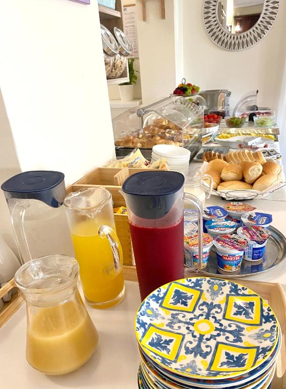 Brokastu galds ar dzērieniem un dažādām uzkodām, tostarp jogurtiem, maizēm un augļiem. Krāsainas šķīvju kaudzes, kas gatavas pasniegšanai.