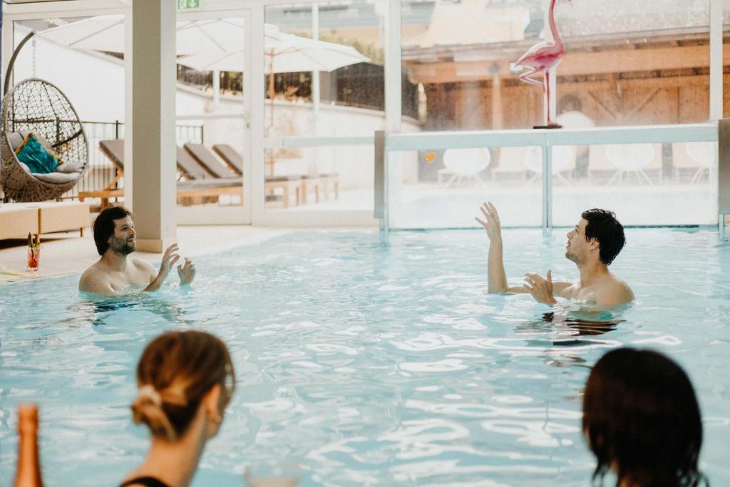 Divi vīrieši sarunājas un izklaidējas baseinā, kamēr sieviete atrodas tuvumā. Fons veido relaksējošu un modernu atmosfēru ar ērtām mēbelēm un saules gaismu.