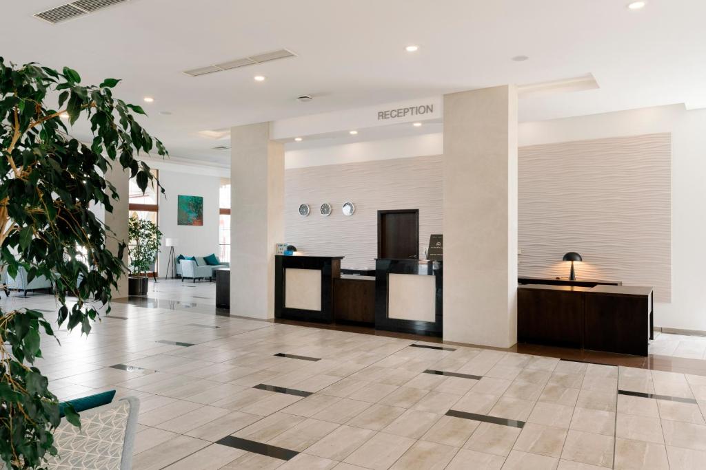 Mūsdienīgs viesnīcas reģistratūras interjers ar elegantu dizainu, kurā redzama recepcija un plaša atpūtas zona ar augiem un dīvāniem.