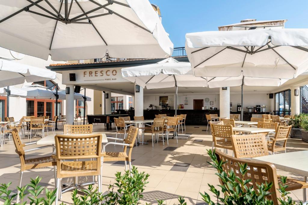 Atvērta terase ar baltiem lietussargiem un koka galdiem, kas ierīkota pie bāra "Fresco". Gaisīga atmosfēra un saulaina diena.
