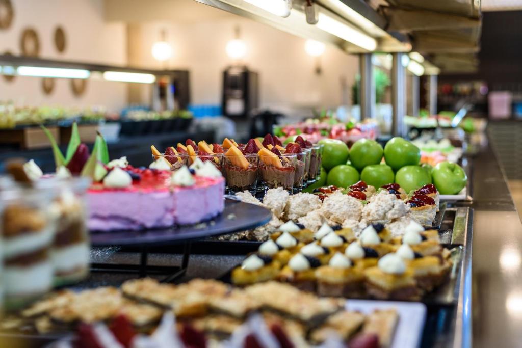 Deserts un augļi krāšņā bufetē, kurā redzami dažādi kūkas, saldējumi un zaļie āboli. Saldumu un augļu daudzveidība rada pievilcīgu un garšīgu atmosfēru.