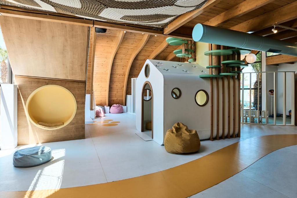 Radoša bērnu spēļu telpa ar koka elementiem, krāsainām sēdvietām un rotaļu mājiņu. Telpa ir gaiša un draudzīga, veidota, lai veicinātu radošumu un iztēli.