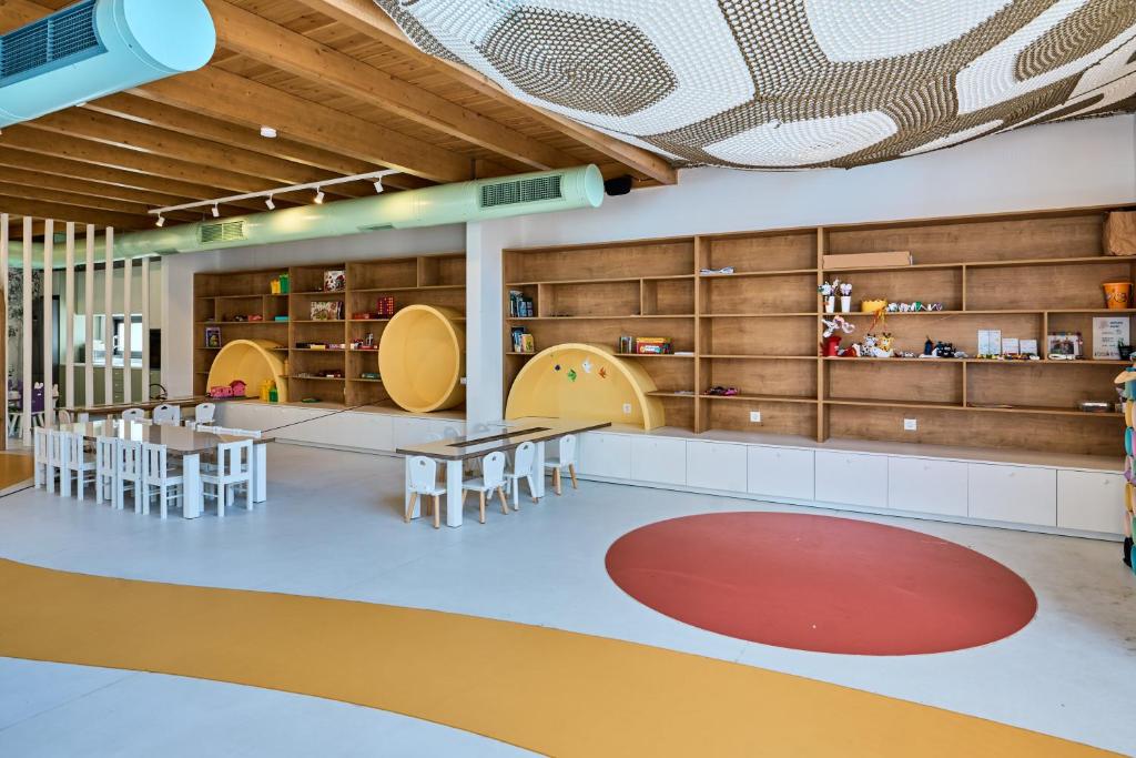 Mūsdienīga bērnu spēļu telpa ar koka griestiem, krāsainām grīdām un plauktiem, kas piepildīti ar rotaļlietām un grāmatām. Telpa ir izgaismota ar dabīgu apgaismojumu, radot draudzīgu un radošu atmosfēru.