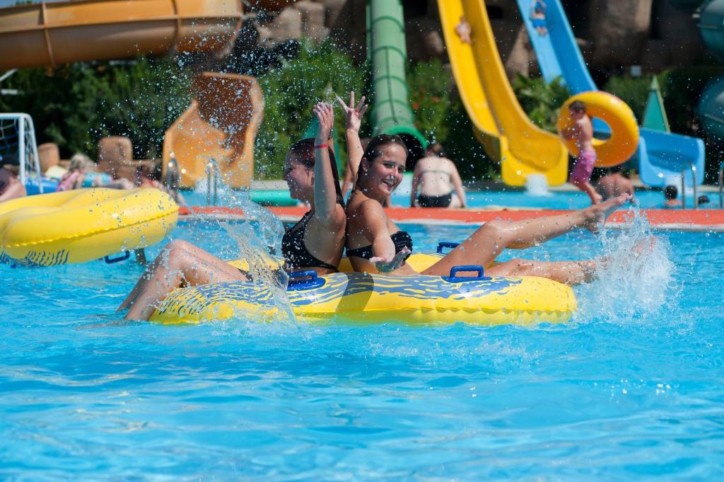 Divas meitenes izbauda ūdens atrakcijas uz dzelteniem plostiem baseinā, izsistot ūdens šļakatas. Fons ir piepildīts ar ūdens slidkalniņiem un apmeklētājiem.