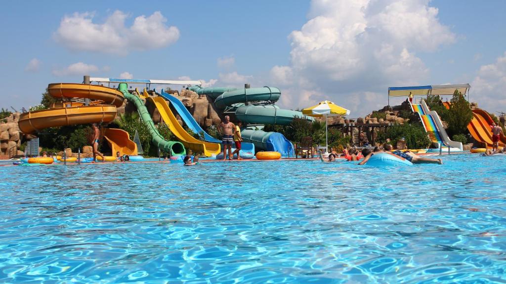 Ūdens atrakciju parks ar vairākiem slidkalniņiem un apmeklētājiem peldbaseinā, kas izbauda saulainu dienu. Skats uz zilo ūdeni un baltiem mākoņiem debesīs.