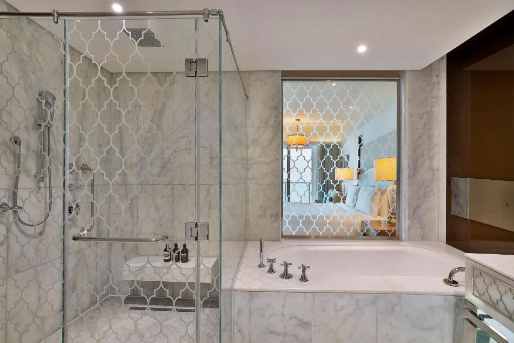 Mūsdienīgs vannas istabas interjers ar stikla dušu un elegantu vannu, kas izceļ marmora sienas un stilīgu dizainu. Redzams arī viesnīcas numura atspulgs.
