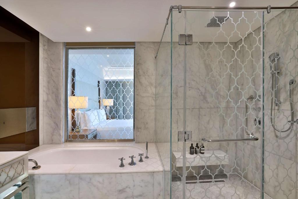 Mūsdienīgs vannas istabas interjers ar marmora sienām, stikla dušu un vannu, kas piedāvā elegantu un relaksējošu atmosfēru. Skats uz guļamistabu caur dekoratīvām stikla durvīm.