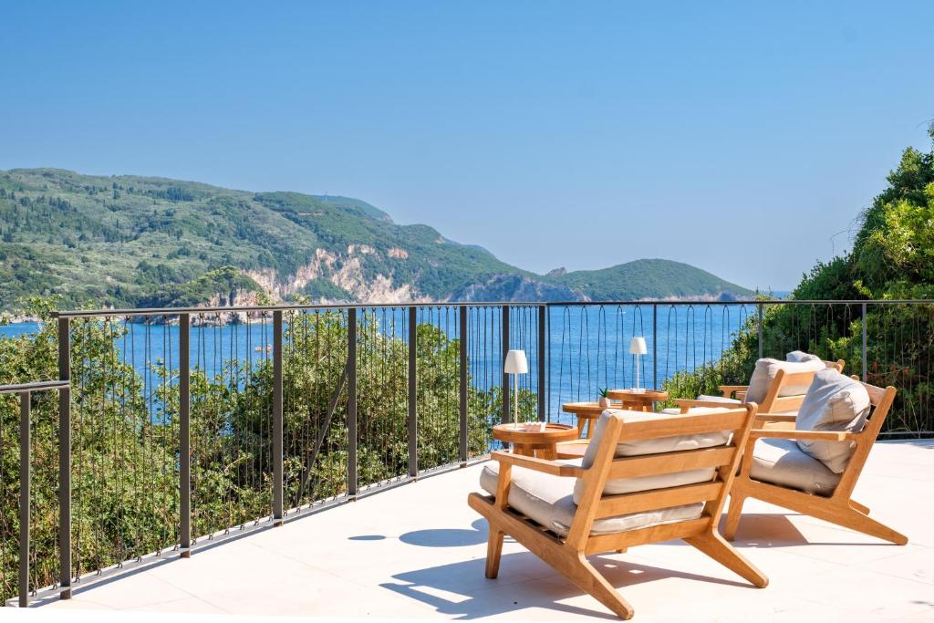 Terase ar skatu uz jūru, kurā redzami divi koka krēsli ar spilveniem un nelieliem galdiņiem. Fons ietver zaļus kalnus un mierīgu ūdeni, kas aicina uz atpūtu.