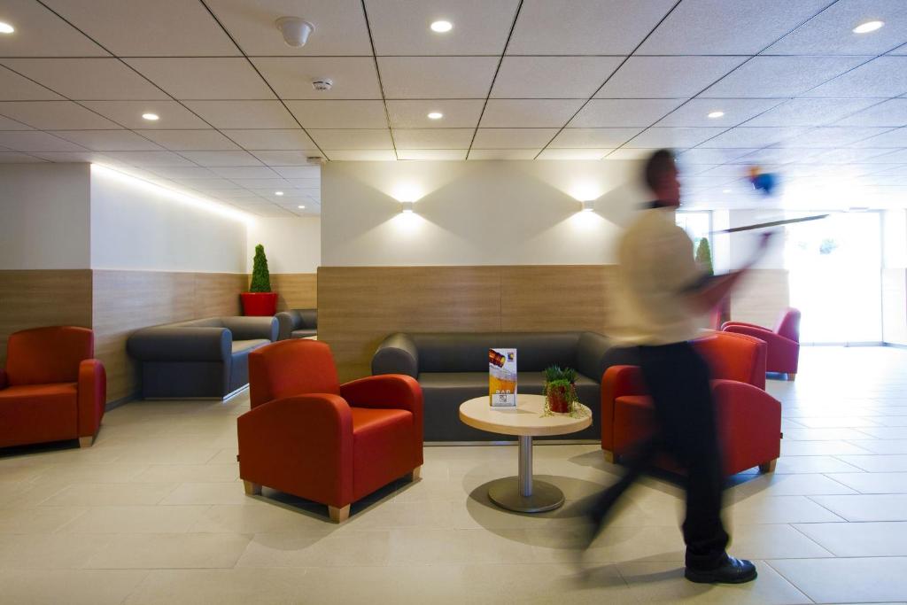 Mūsdienīgs viesnīcas hols ar sarkanām un pelēkām mēbelēm, apgaismojums un zaļiem augiem. Redzams darbinieks, kas pārvietojas pa telpu.