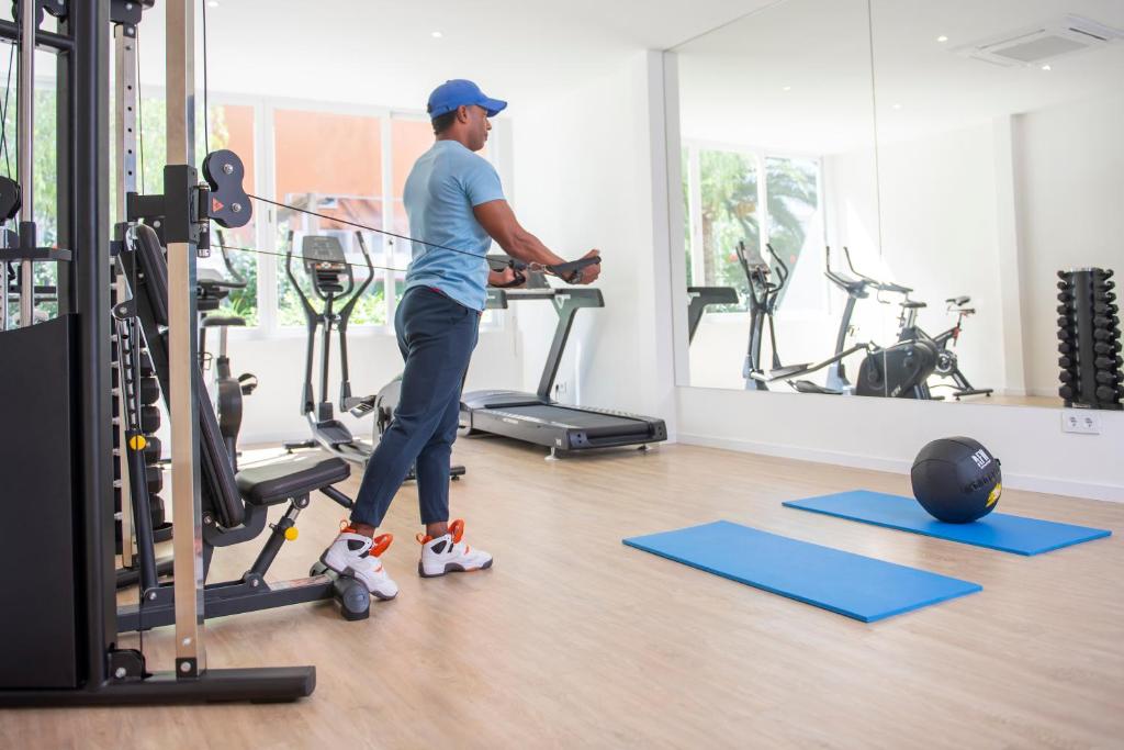 Vīrietis trenējas sporta zālē, izmantojot trenažieri ar stiegru, kamēr blakus ir skrejceliņš un sporta inventārs. Telpa ir gaiša un moderna, ar lielām logu stiklu virsmām.