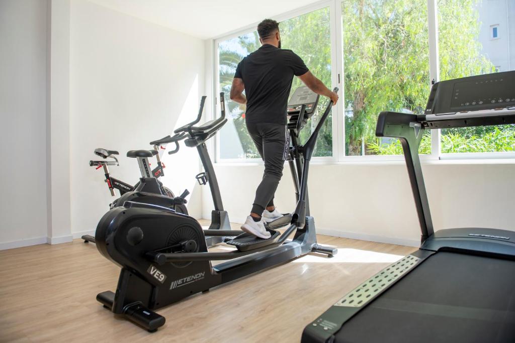 Vīrietis vingro uz eliptiskā trenažiera modernā sporta zālē ar lieliem logiem, kas piedāvā dabīgu apgaismojumu un skatu uz zaļumiem. Blakus redzami divi velosipēdi un skrejceļš.