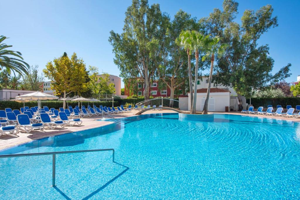 Liels peldbaseins ar zilu ūdeni, apjozts ar sauļošanās krēsliem un palmu kokiem, kas rada relaksējošu atmosfēru. Apkārtējā ainava ir zaļa un saulaina, ideāla atpūtai.