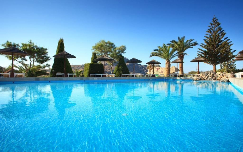 Zilā baseina ainava ar palmām un saulessargiem, kas veido relaksējošu atmosfēru. Skaidrs debesis un saules gaisma papildina tropisko noskaņu.