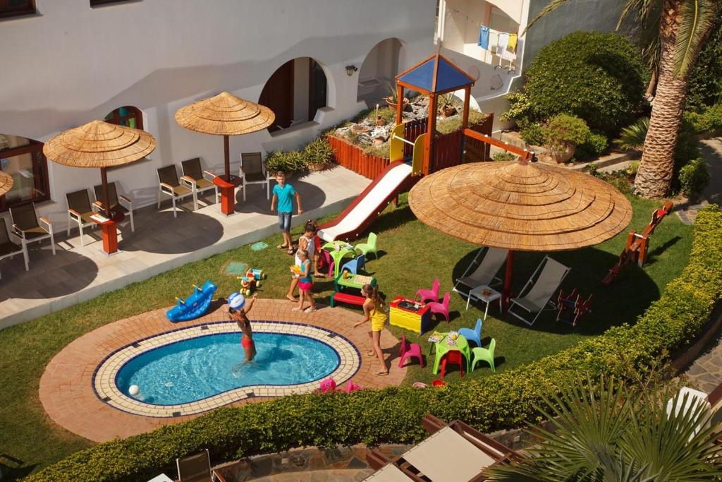 Bērnu rotaļu laukums ar mazu baseinu, saulessargiem un slidkalniņu, kur bērni izbauda ūdens priekus un spēlējas. Vasarīgs skats uz viesnīcas dārzu ar krāsainām bērnu mēbelēm.