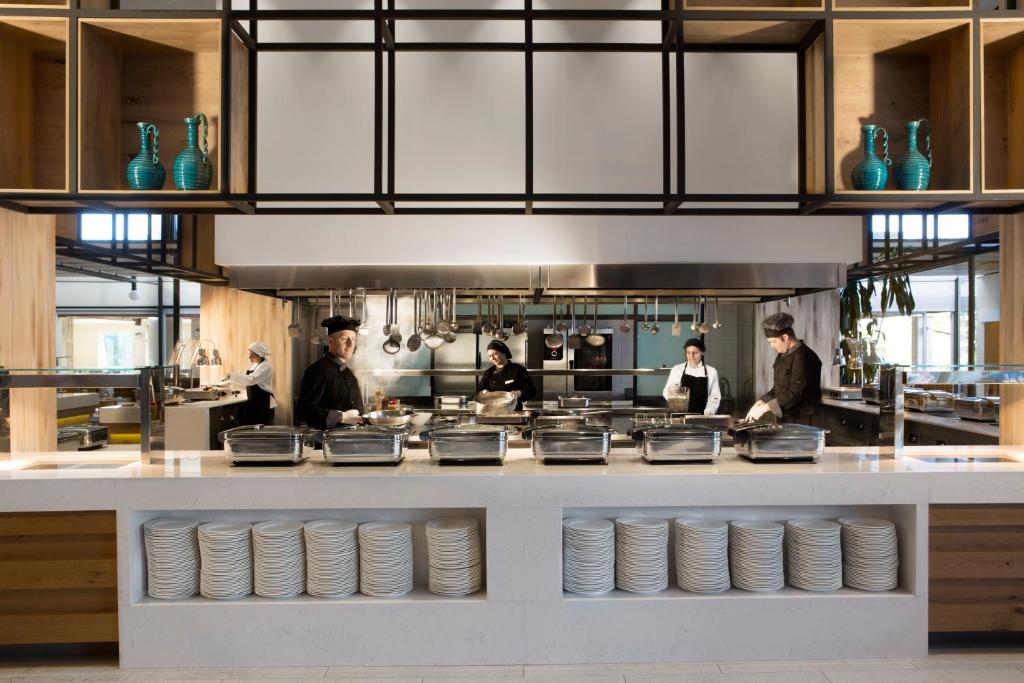 Mūsdienīga virtuve ar šefpavāriem, kas gatavo ēdienus, un elegantu interjeru, kurā redzami stikla trauki un tīras baltas šķīvju rindas.