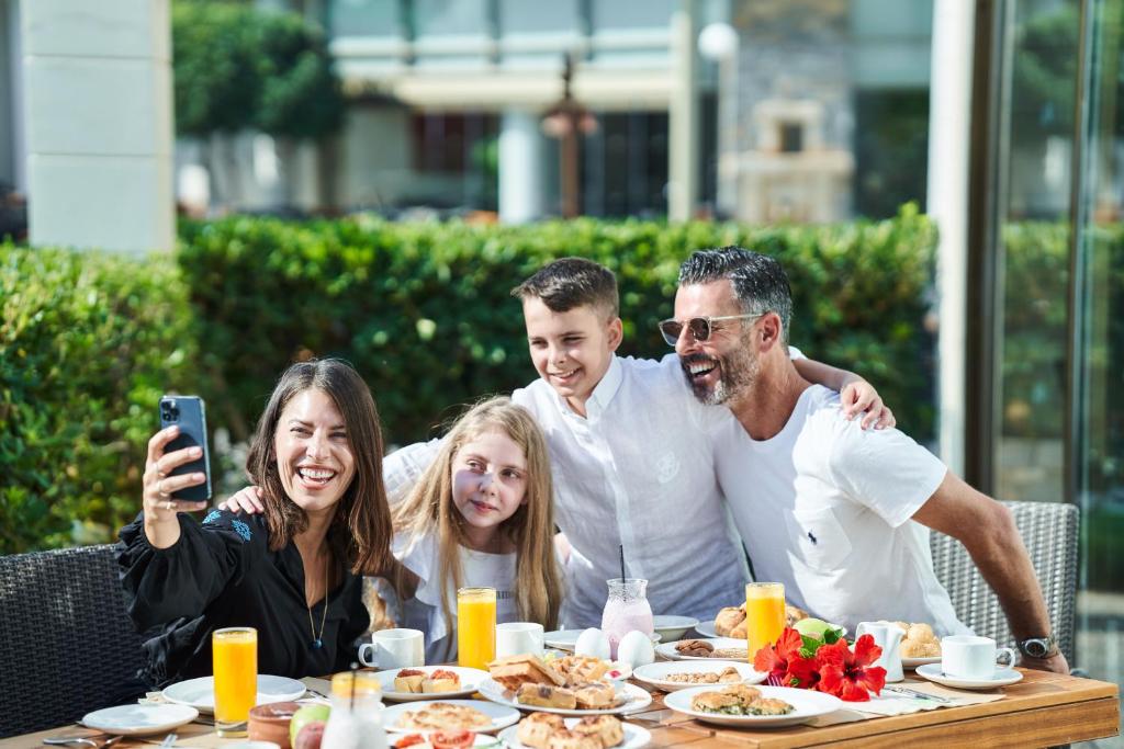 Ģimene kopā priecīgi fotografējas pie galda ar brokastīm, baudot rīta sauli un gardus ēdienus. Galda klājumā redzami daudzi dzērieni un uzkodas.