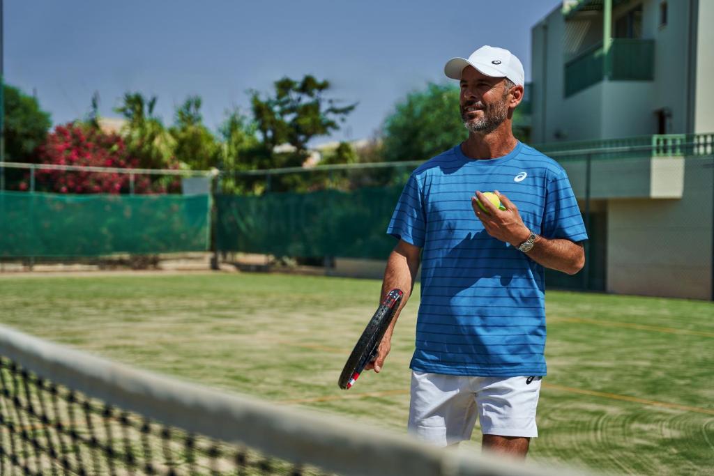 Vīrietis, tērpies zilo tenisa krekli un baltās šortos, tur tenisa raketi un bumbu, stāvot uz tenisa korta ar zaļu fona.