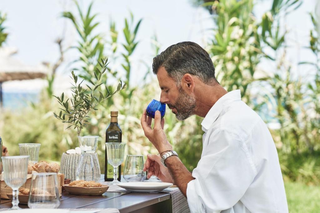 Vīrietis baltā kreklā sēž pie galda ārā, baudot dzērienu no zilas krūzes, blakus ir ēdieni un olīveļļa. Apkārt ir zaļumi un pludmales skats.
