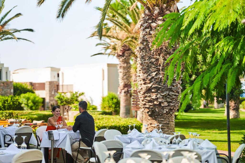 Romantiska pusdienu ainas ārpusē ar palmām un zaļu dārzu. Pāris, apģērbts eleganti, sēž pie galda ar vīna glāzēm, baudot laiku kopā.