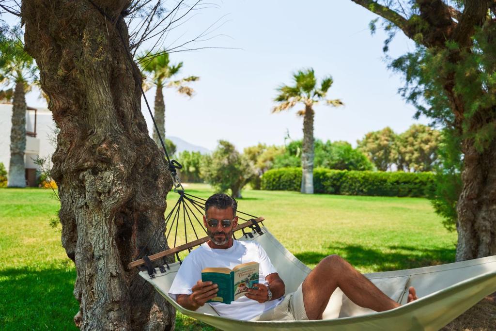 Vīrietis atpūšas šūpuļtīklā zem palmu kokiem, lasot grāmatu saulainā dārzā. Apkārt ir zaļa zāle un ainaviskas ainavas.
