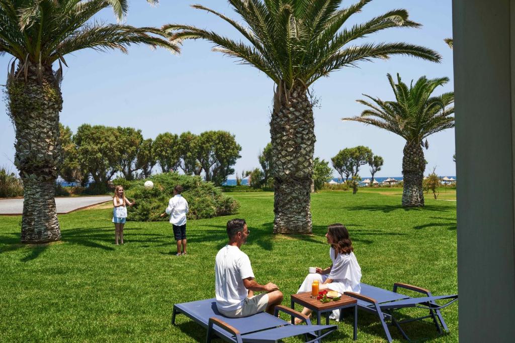 Atpūtas ainava ar palmām, kur divi cilvēki sēž uz sauļošanās krēsliem, baudot dzērienus un uzkodas, kamēr divi bērni spēlējas ar bumbu zaļajā zālē. Fons ietver jūru un pludmales lietas.