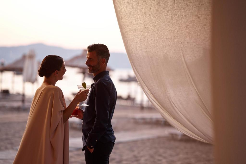 Pāris, kas stāv pludmalē un smaidīgi sarunājas, turot kokteiļus rokās. Aiz viņiem ir smilšu pludmale un saulrieta skats.